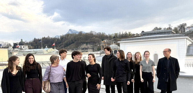 A group of people stands on a rooftop overlooking an urban landscape. They are dressed in elegant, dark clothing and appear cheerful in their shared environment. | © Universität Mozarteum