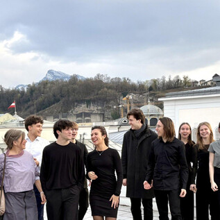 Un groupe de personnes se tient sur un toit avec vue sur un paysage urbain. Ils portent des vêtements élégants et sombres et semblent joyeux dans leur environnement commun. | © Universität Mozarteum