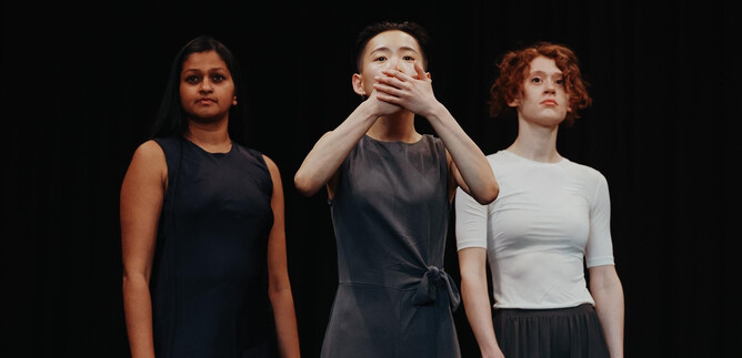 Three women are standing on a stage against a black background. One of the women has her hands in front of her mouth and makes an expressive gesture. | © Jasmin Walter
