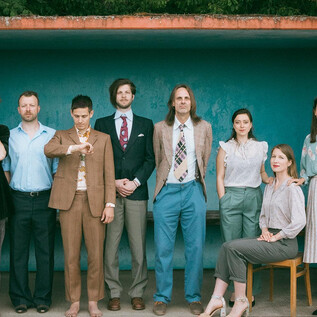 A group of nine people in formal attire stands in front of a colorful background. They are posing in different stances and showcasing a diverse use of colors and styles. | © Sonja Herpich