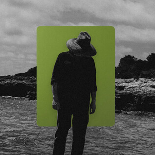 A man stands on the beach with a hat and has his head lowered. In the background, there are waves and a cloudy sky. | © James Keane