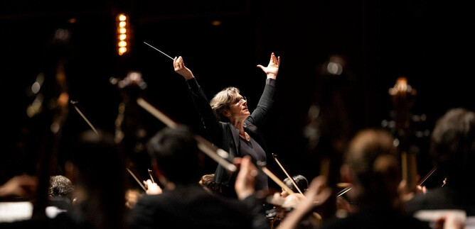 A conductor leads an orchestra with passionate gestures. The musicians play with focus, surrounded by a dark background. | © Erika Mayer
