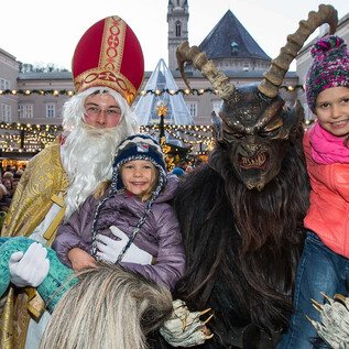 A festive scene with two children dressed up as Santa Claus and a Krampus. In the background, a bustling Christmas market atmosphere can be seen. | © Salzburger Christkindlmarkt