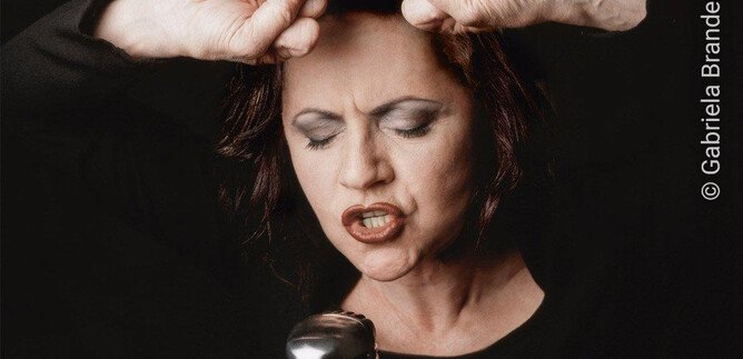 Una cantante que canta apasionadamente en un micrófono. Sus ojos están cerrados y tiene las manos levantadas sobre la cabeza. | © Gabriela Brandenstein