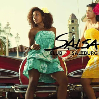 Deux femmes en robes colorées posent devant une voiture classique rouge. En arrière-plan, on peut voir des bâtiments historiques et des palmiers. | © Jazzit_Salsa Club Salzburg