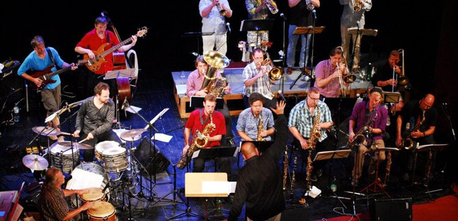 Eine große Band mit vielen Musikern spielt auf einer Bühne. Der Dirigent leitet das Orchester, während verschiedene Instrumente zu hören sind. | © Karoline Muehlbacher