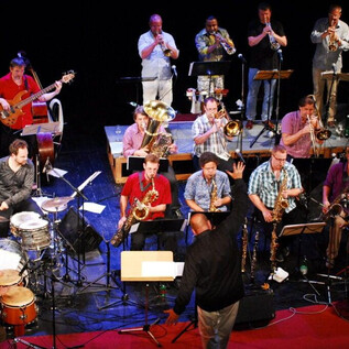 A large band with many musicians is playing on a stage. The conductor is leading the orchestra while different instruments can be heard. | © Karoline Muehlbacher