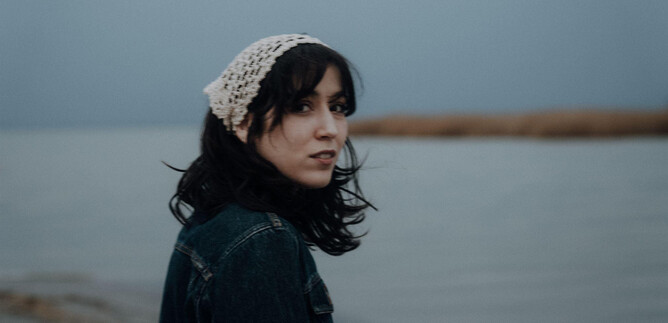 A young woman with dark hair stands by the water. She is wearing a light headscarf and looking at the camera. | © Ines Futterknech