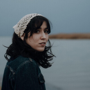 A young woman with dark hair stands by the water. She is wearing a light headscarf and looking at the camera. | © Ines Futterknech