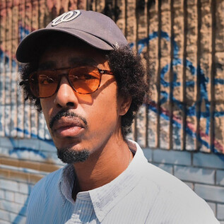 Ein Mann steht vor einer farbenfrohen Wand mit Graffiti. Er trägt eine Kappe und Sonnenbrille und hat lockiges Haar. | © Rockhouse Salzburg