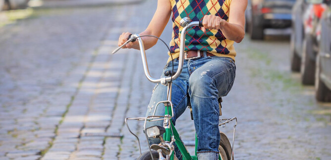 A man rides a green bicycle along the cobbled street. The surroundings are urban with cars parked at the side of the road. | © Amac Garbe