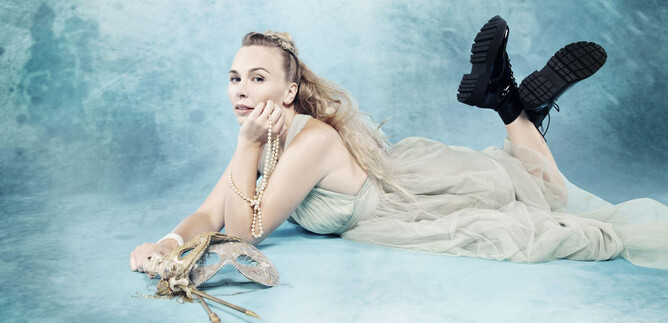 A woman is lying on a blue background. She is wearing a flowing dress and boots while holding elegant accessories in her hand. | © Simon Fowler