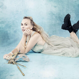 A woman is lying on a blue background. She is wearing a flowing dress and boots while holding elegant accessories in her hand. | © Simon Fowler