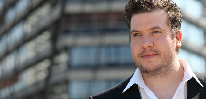 Un uomo in un completo sta di fronte a un edificio moderno. Ha un'espressione sicura di sé e guarda nella telecamera. | © christian-kargl