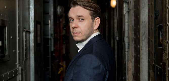 A man in a suit is looking over his shoulder in a narrow, illuminated corridor. The background is dark and industrial. | © Edouard Brane