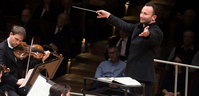 An orchestra conductor passionately leads a performance with expressive gestures. In the foreground, a violinist plays while the audience listens attentively. | © Monika Rittershaus