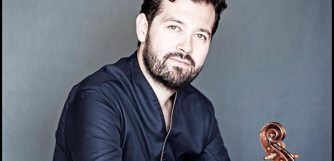 Un uomo con la barba è seduto davanti a uno sfondo neutro. Tiene un violino in mano e guarda direttamente nella telecamera. | © Giorgia Bertazzi