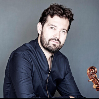 A man with a beard is sitting in front of a neutral background. He is holding a violin in his hand and looking directly at the camera. | © Giorgia Bertazzi