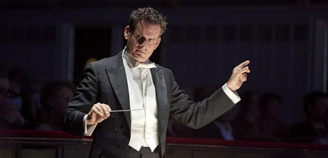 Un direttore d'orchestra guida un'orchestra con grande passione. Sullo sfondo si possono vedere spettatori durante un concerto. | © Michael Poehn