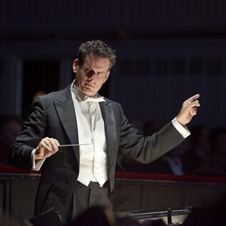 A conductor leads an orchestra with great passion. In the background, spectators can be seen during a concert. | © Michael Poehn