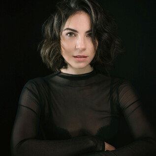 A woman with short, curly hair is wearing a black, transparent top. She is looking directly into the camera against a dark background. | © Chris Gonz