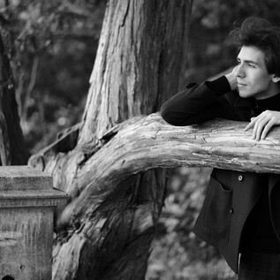 A thoughtful young man leans against a tree trunk outdoors. The image composition is in black and white. | © Sasha Gusov