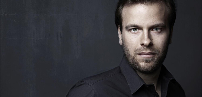 A portrait of a young man with short dark hair and a beard. He is wearing a black shirt and looking directly at the camera. | © Gisela Schenker