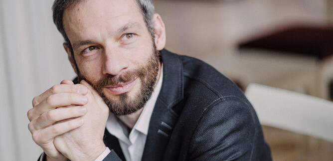 A man with short hair and a beard sits thoughtfully. He is wearing a black suit and looking kindly at the camera. | © Marco Borggreve