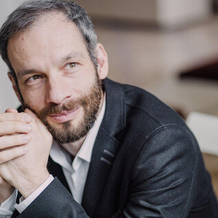 Un homme aux cheveux courts et à la barbe est assis pensif. Il porte un costume noir et regarde amicalement l'appareil photo. | © Marco Borggreve