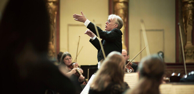 Ein Dirigent leitet ein Orchester in einem prächtigen Saal. Musiker aus verschiedenen Instrumentengruppen konzentrieren sich auf seine Anweisungen. | © Meinrad Hofer