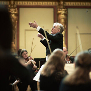 Un chef d'orchestre dirige un orchestre dans une salle magnifique.  
Des musiciens de différents groupes d'instruments se concentrent sur ses instructions. | © Meinrad Hofer