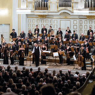An orchestra in full force is playing on a stage. The audience in the background is applauding enthusiastically. | © Albert Moser