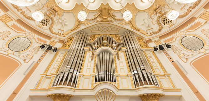 A large, magnificent organ with many pipes in a festively designed room. The walls are painted in soft colors and adorned with golden embellishments. | © Wolfgang Lienbacher