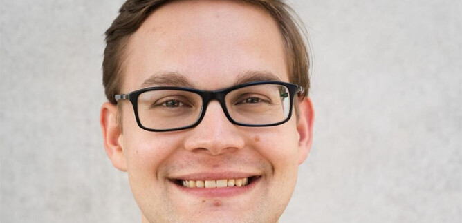 Un giovane uomo con occhiali sorride alla telecamera. Lo sfondo è chiaro e neutro. | © www.salzburger-dom.at