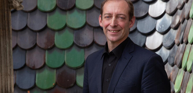 Un uomo sorride davanti a una facciata colorata di scandole. Indossa un abito scuro e ha un'espressione amichevole. | © Verein "Unser Stephansdom"