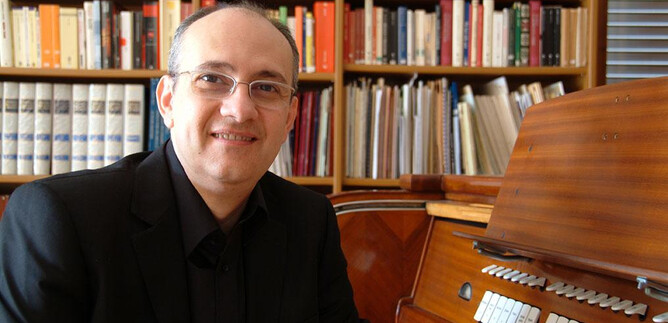 Un uomo sorride seduto accanto a uno strumento musicale in una stanza con librerie sullo sfondo. Indossa una camicia nera e degli occhiali. | © Roberto Marini