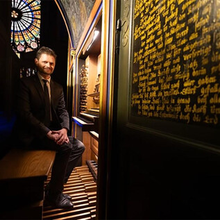 A man sits in a historic room with an organ. In the background, colorful stained glass windows and golden writing on the wall can be seen. | © Jérôme Dorkel - Ville de Strasbourg