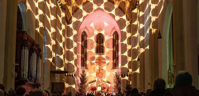 A festively illuminated interior of a church with an audience group. Mood-enhancing light patterns fall from the ceiling, creating a special atmosphere. | © Original Salzburger Advent