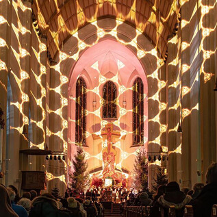 A festively illuminated interior of a church with an audience group. Mood-enhancing light patterns fall from the ceiling, creating a special atmosphere. | © Original Salzburger Advent