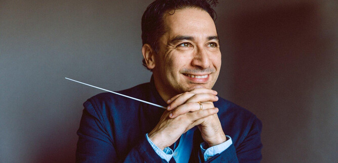 A smiling man with a staff in his hand sits thoughtfully. He is wearing a blue suit and has a friendly expression. | © Julia Sellmann