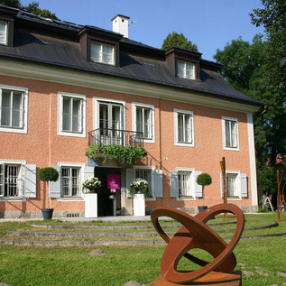 Ein schönes, rosa Haus mit weißen Fenstern und einem Balkon. Im Vordergrund steht eine moderne Skulptur auf einer grünen Wiese. | © P. Raas