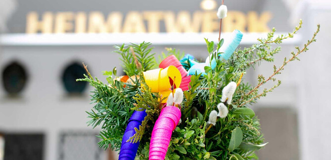 Egy színes csokor zöld növényekkel és színes, spirális díszítésekkel. A háttérben egy tábla látható, rajta a "HEIMATWERK" felirattal. | © Salzburger Heimatwerk