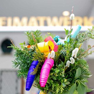 Ein bunter Strauß mit grünen Pflanzen und bunten, spiralförmigen Dekorationen. Im Hintergrund ist ein Schild mit der Aufschrift "HEIMATWERK" zu sehen. | © Salzburger Heimatwerk
