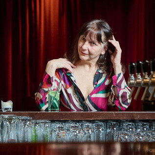 Eine Frau sitzt an einer Bar und schaut aufmerksam. Im Hintergrund sind Gläser und eine Theke mit Zapfhähnen zu sehen. | © Foto Flausen