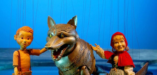 A boy doll, a girl doll in a red cloak, and a wolf doll stand together. The background is blue, creating an imaginative atmosphere. | © Salzburger Marionettentheater