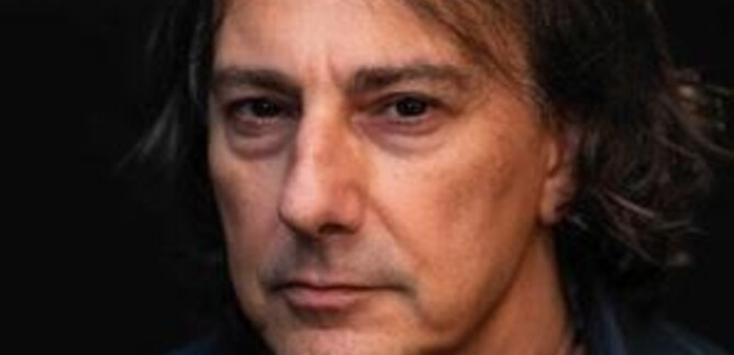 A man with long, dark hair and a serious expression stands in front of a dark background. He is wearing a black shirt and a pearl necklace. | © derkonzertfotograf