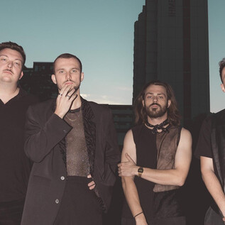 Egy négy férfiból álló csoport egy városi háttér előtt áll. A jelenet modern és elegáns hangulatot sugall. | © Arne Müseler