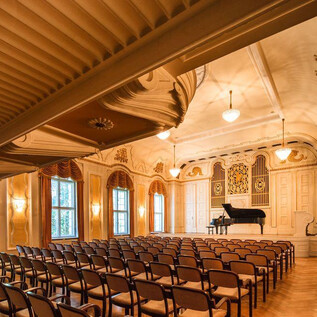 Un elegante salón de conciertos con un escenario y un piano. Las filas de sillas en el frente están listas para los invitados. | © Christian Schneider