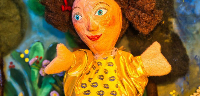 A colorful doll with curly hair and yellow clothing. In the background, cheerful colors and a fairy tale landscape can be seen. | © Hannes Öhlböck