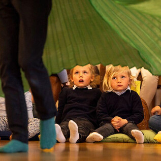 Two children are sitting excitedly on the floor watching a performance. In the background, a colorful curtain can be seen. | © Erika Mayer